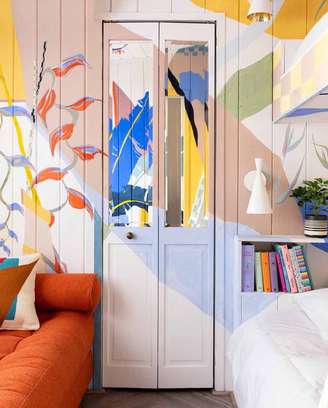 Stylish bedroom interior featuring a colorful mural, painted white double doors, orange sofa, and decorative pillows, creating a vibrant space.