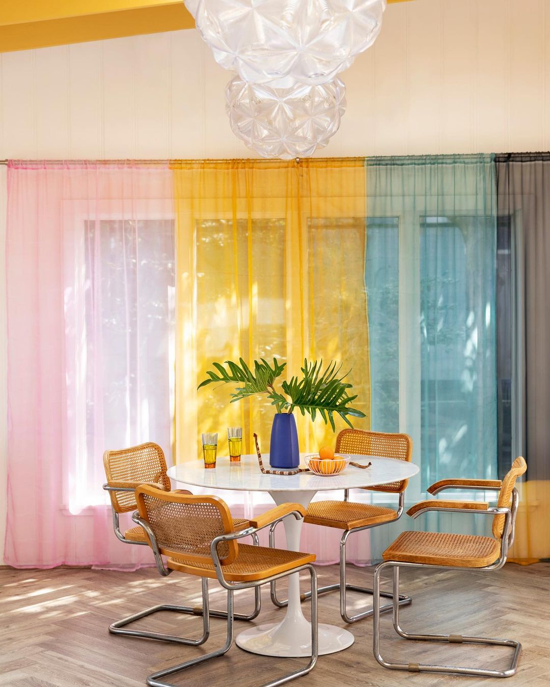 Dining room interior with modern tulip table, rattan chairs, colorful sheer curtains, and decorative chandelier, creating a bright, airy aesthetic.