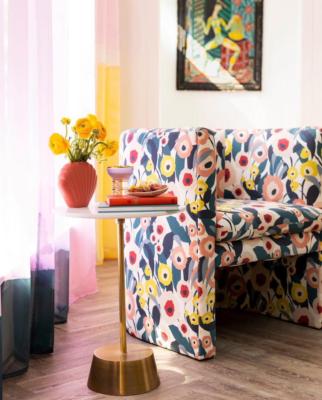 Interior corner of a room with a floral pattern chair, accent table holding flowers and snacks, and bright, sheer curtains.