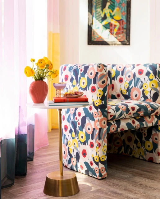 Interior corner of a room with a floral pattern chair, accent table holding flowers and snacks, and bright, sheer curtains.