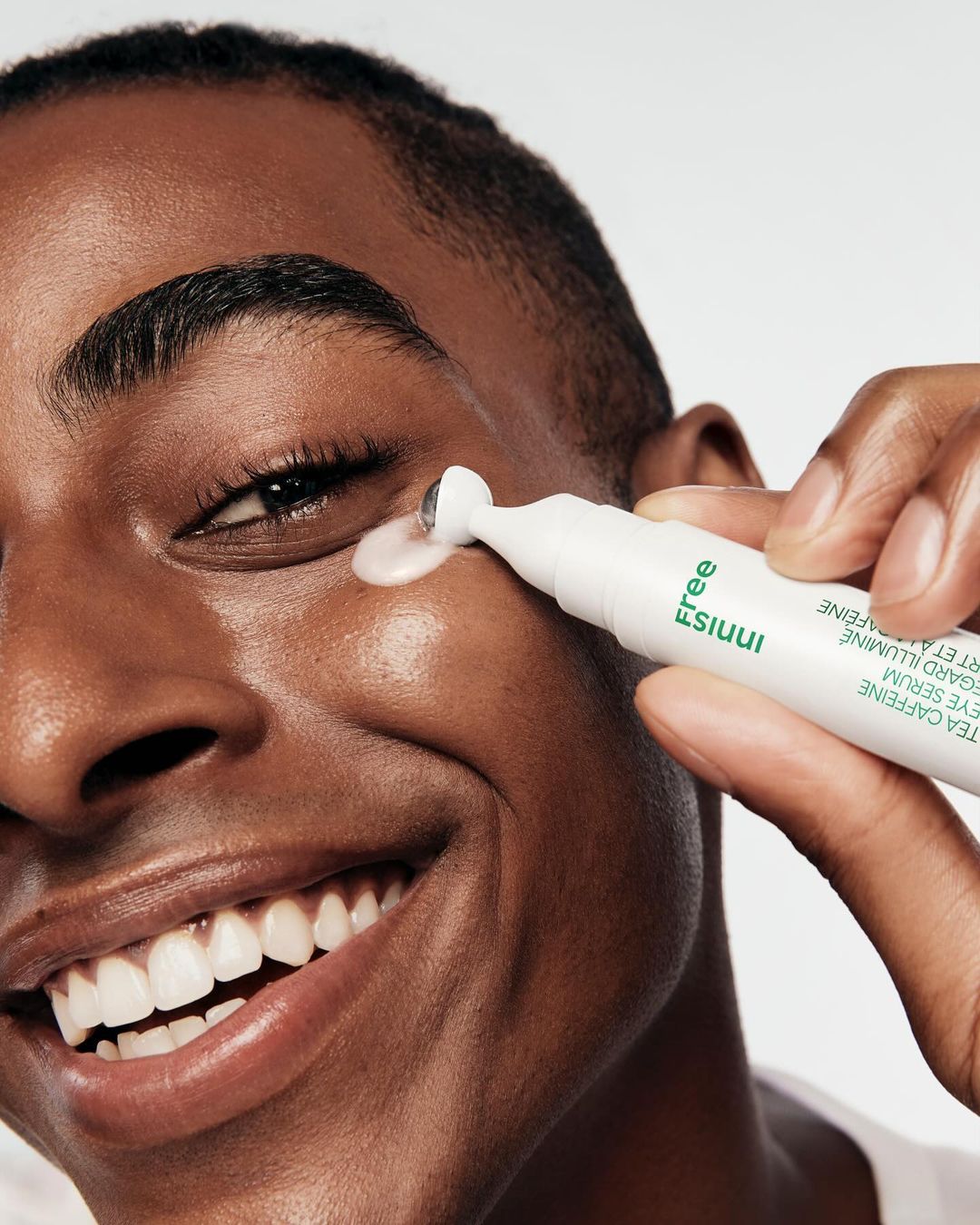 Close-up of a smiling, dark-skinned man applying an eye serum roller to his eye. Product is white tube with green text.