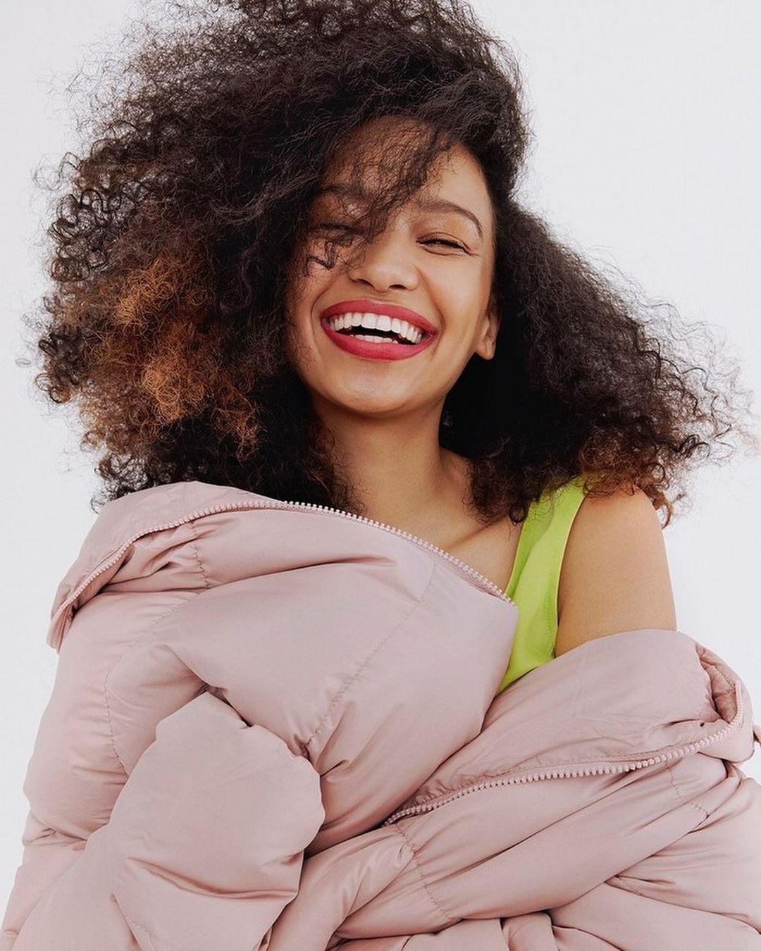 Portrait of smiling mixed-race woman with curly hair in a photo studio, wrapped in a pink puffer jacket and wearing a chartreuse tank top.