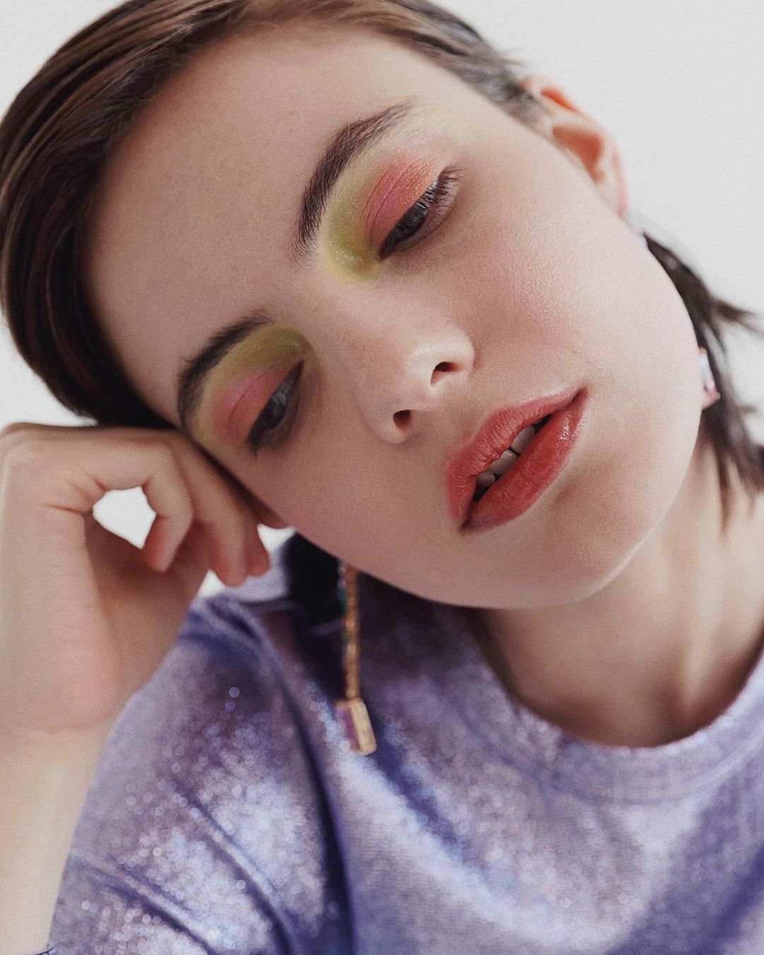 Close-up portrait of a young woman with colorful eyeshadow and shimmery lavender top in a bright studio setting with a neutral backdrop.