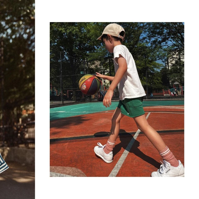 Young boy in shorts and cap dribbles a colorful basketball on an outdoor court surrounded by trees and city buildings in the background.