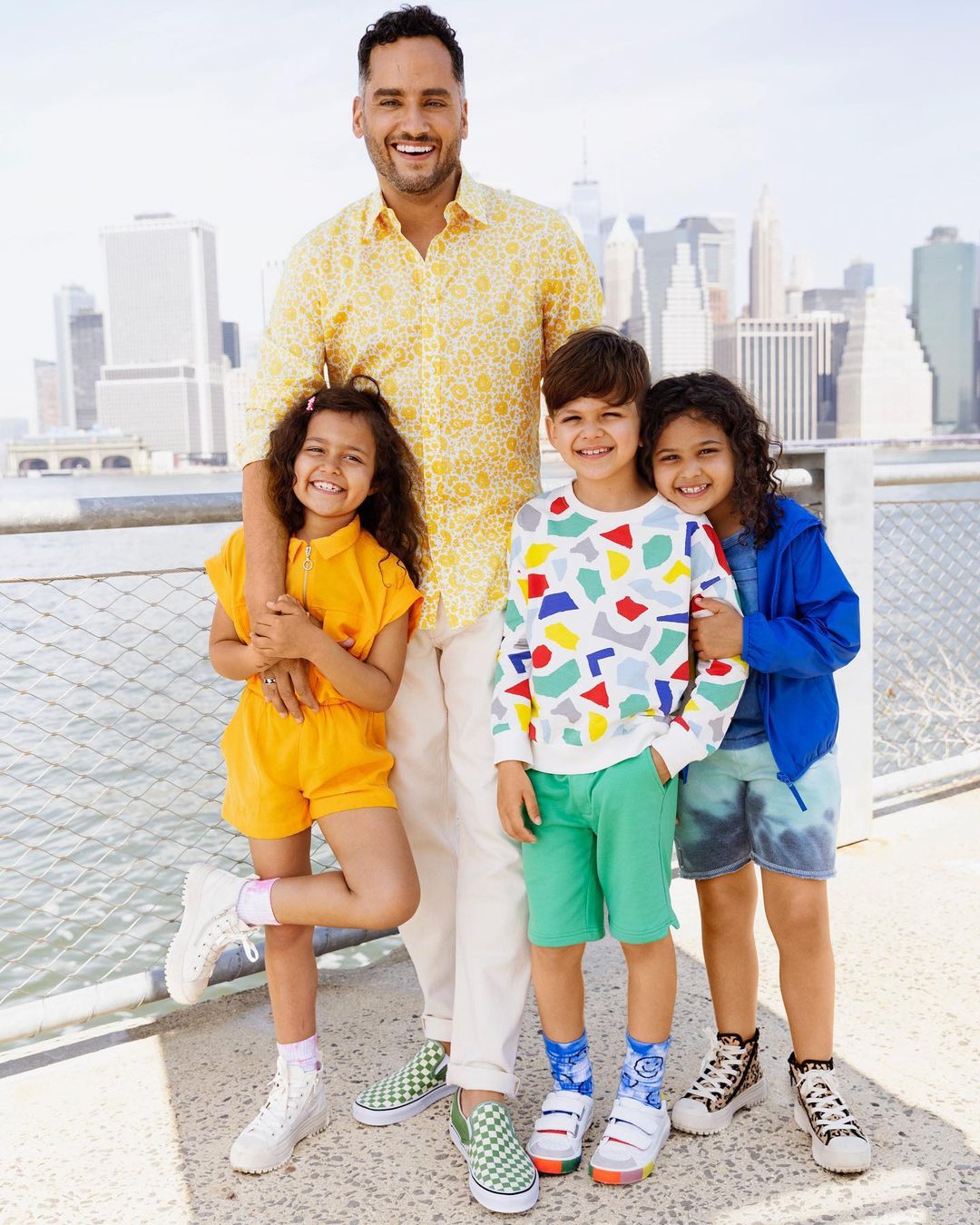 Happy family of four poses outdoors with city buildings in background. Father in patterned shirt stands with three smiling children.