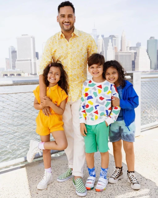 Happy family of four poses outdoors with city buildings in background. Father in patterned shirt stands with three smiling children.