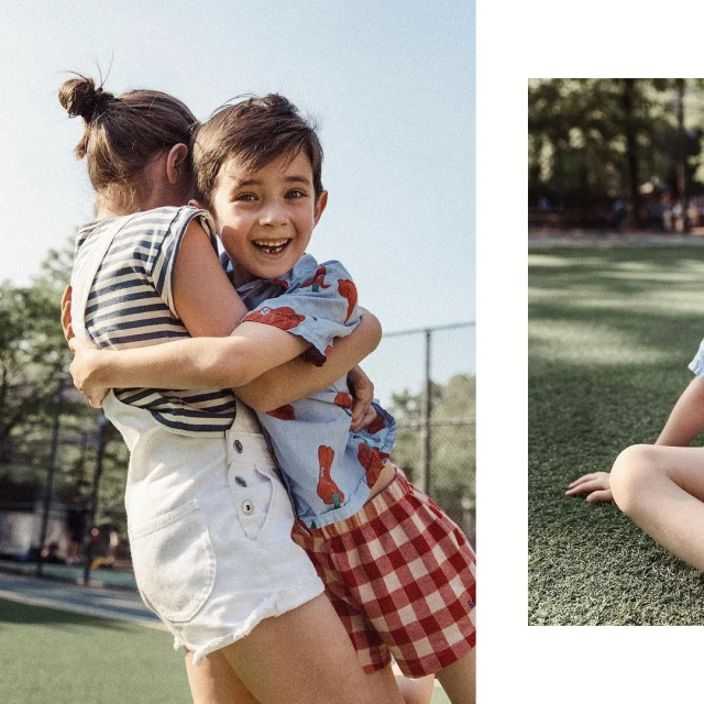 Two children, a boy and a girl, are outdoors embracing. The boy smiles, wearing a patterned shirt and red plaid shorts. The girl wears overalls.