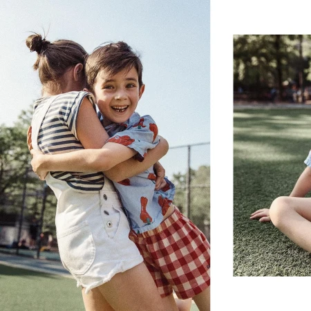 Two children, a boy and a girl, are outdoors embracing. The boy smiles, wearing a patterned shirt and red plaid shorts. The girl wears overalls.