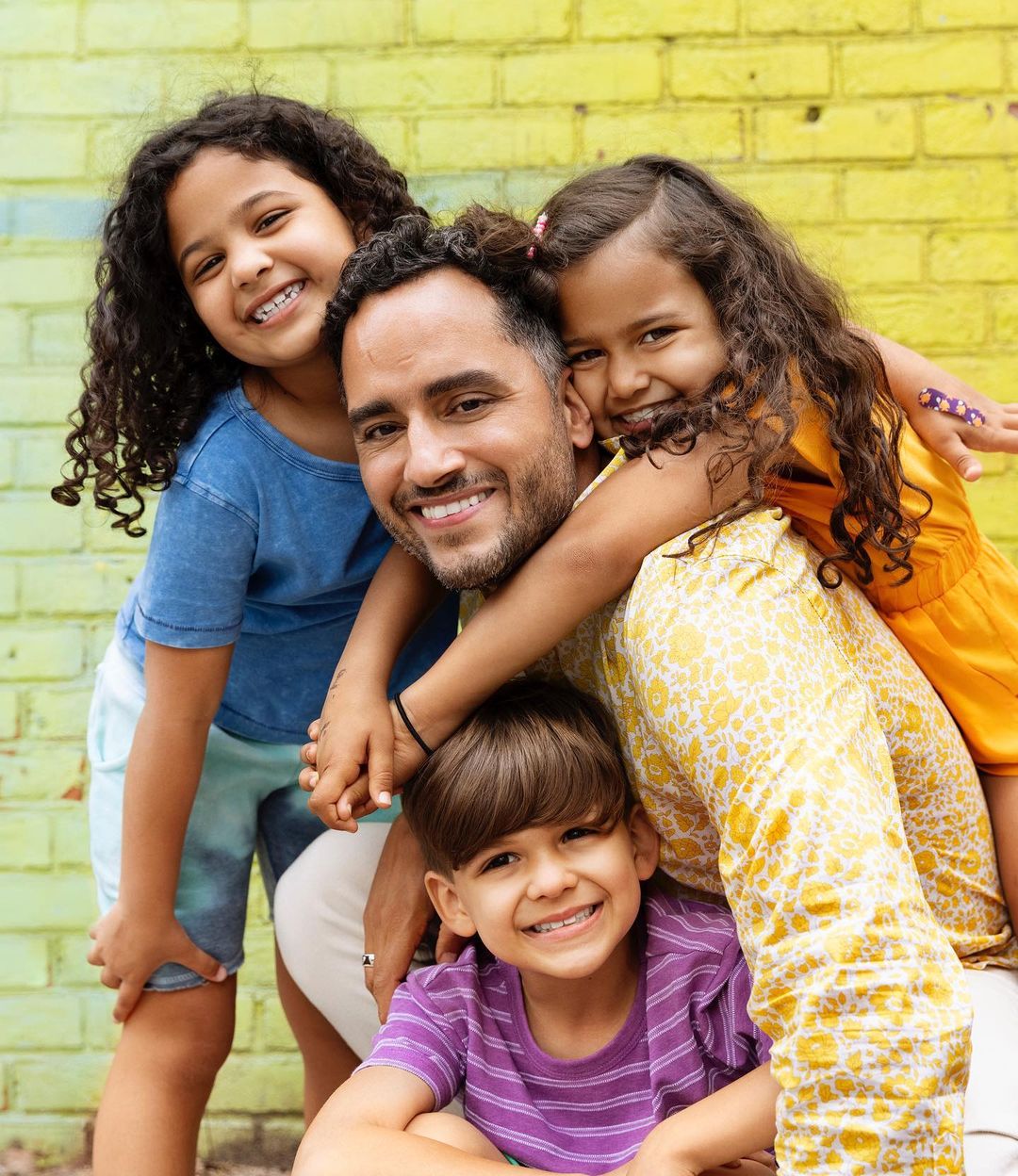 Smiling father hugs three kids against bright, colorful brick wall outdoors. A happy family portrait with natural lighting.