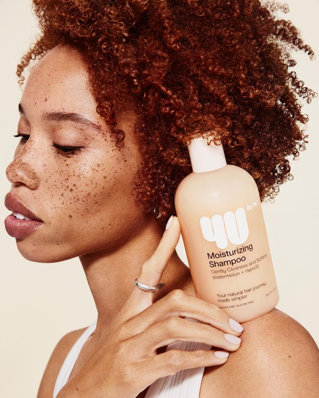Woman with curly hair holding a bottle of moisturizing shampoo, showcasing beauty and hair care product in a light-toned setting.