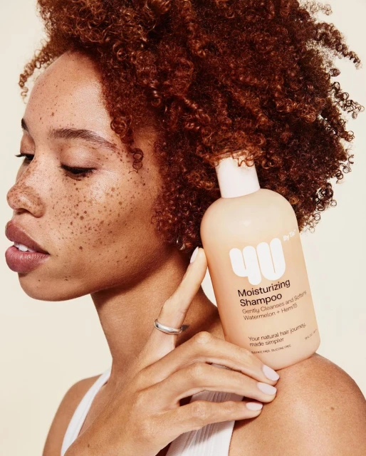 Woman with curly hair holding a bottle of moisturizing shampoo, showcasing beauty and hair care product in a light-toned setting.