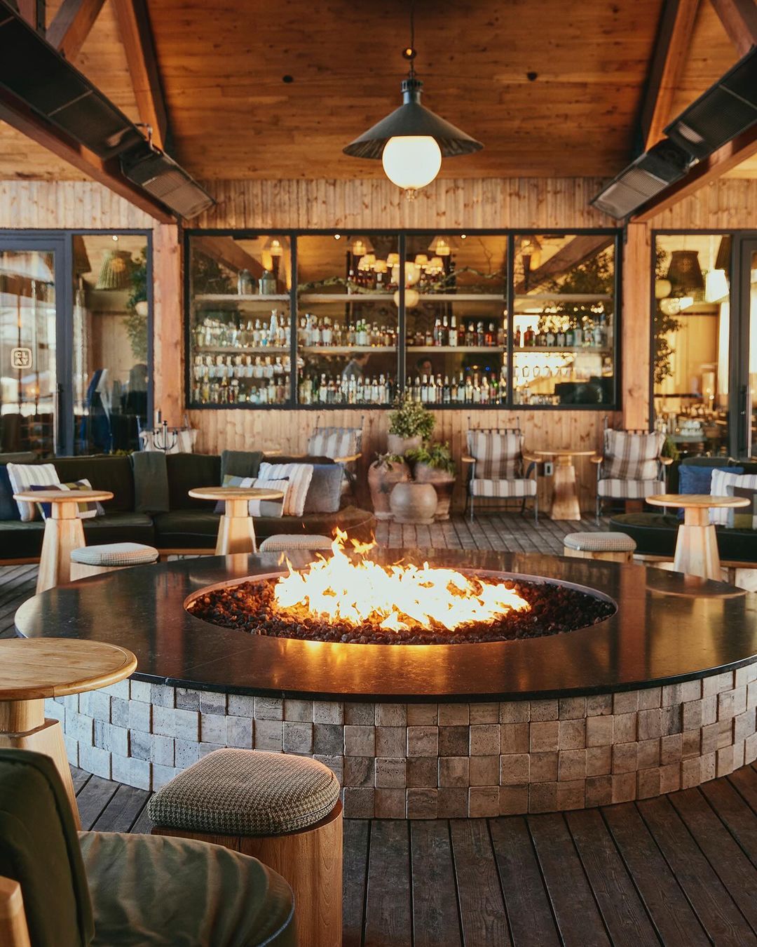 Indoor lounge area featuring a circular stone fire pit with flames, surrounded by seating, wooden floors, and a stocked bar display.