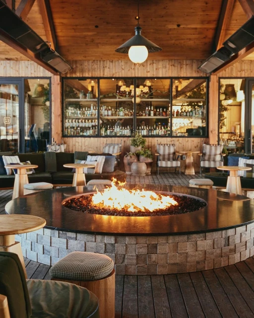 Indoor lounge area featuring a circular stone fire pit with flames, surrounded by seating, wooden floors, and a stocked bar display.