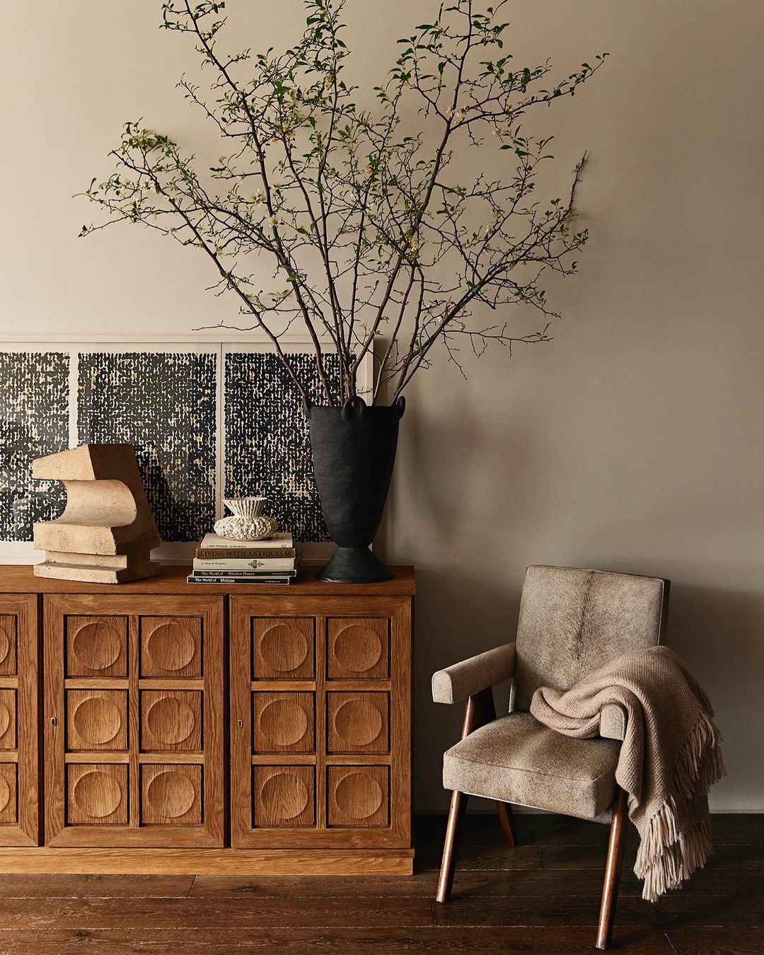 Living room interior featuring a wooden cabinet with a decorative vase, bare branches, stacked books and an armchair with a throw blanket.