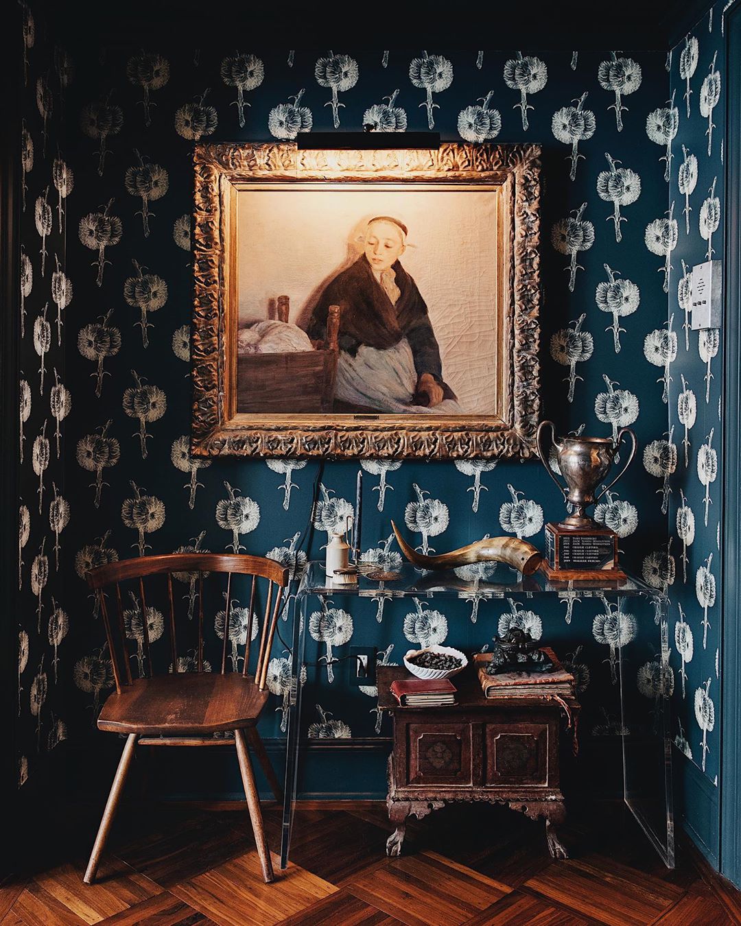 Interior shot of an antique-style room featuring a framed painting above a glass table adorned with vintage objects and a wooden chair.