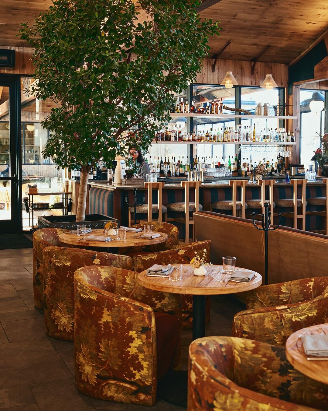 Stylish restaurant interior featuring warm wood tones, round tables with patterned chairs, and a bar area with bottles and glasses.