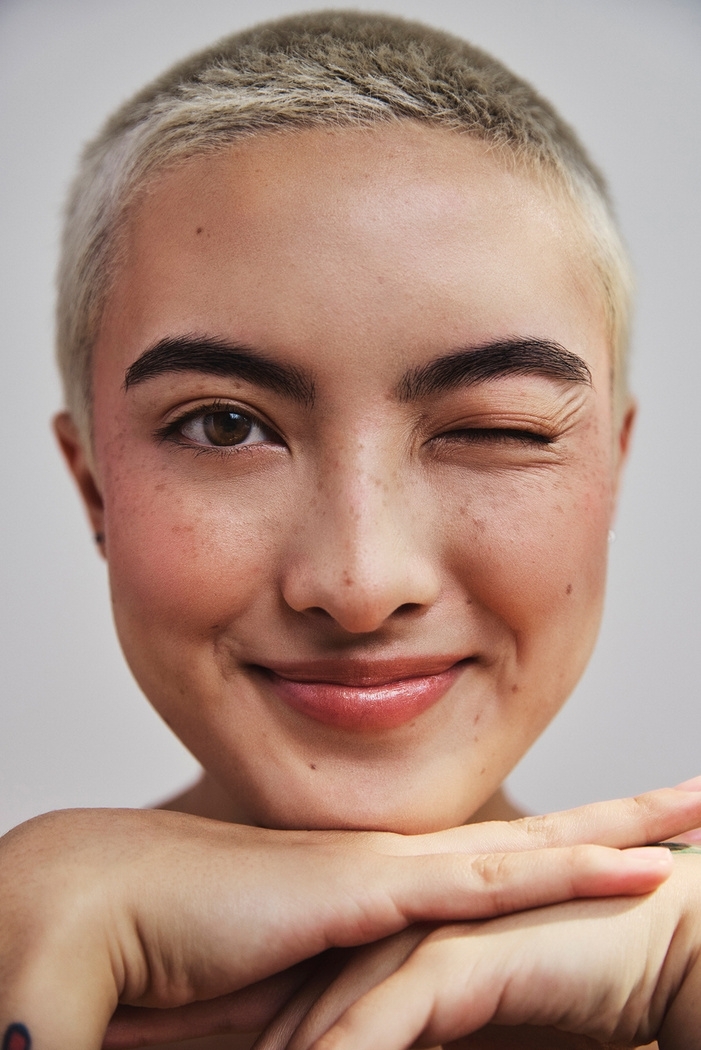 Close-up portrait of a smiling person with short dyed blonde hair and freckles winking at the camera.