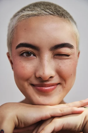 Close-up portrait of a smiling person with short dyed blonde hair and freckles winking at the camera.