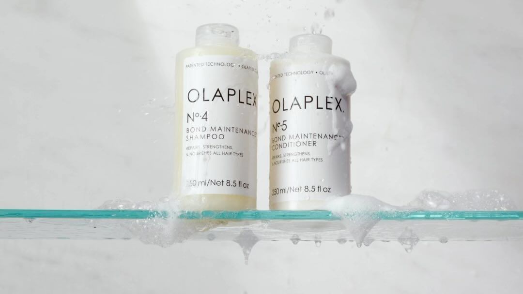 Olaplex shampoo and conditioner bottles on a glass shelf with water splashing against a white background in a beauty product shot.