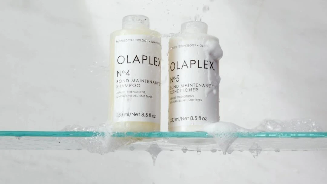 Olaplex shampoo and conditioner bottles on a glass shelf with water splashing against a white background in a beauty product shot.