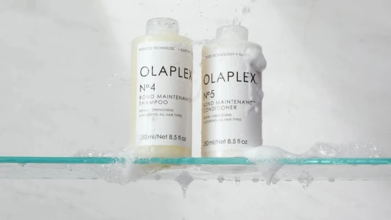 Olaplex shampoo and conditioner bottles on a glass shelf with water splashing against a white background in a beauty product shot.