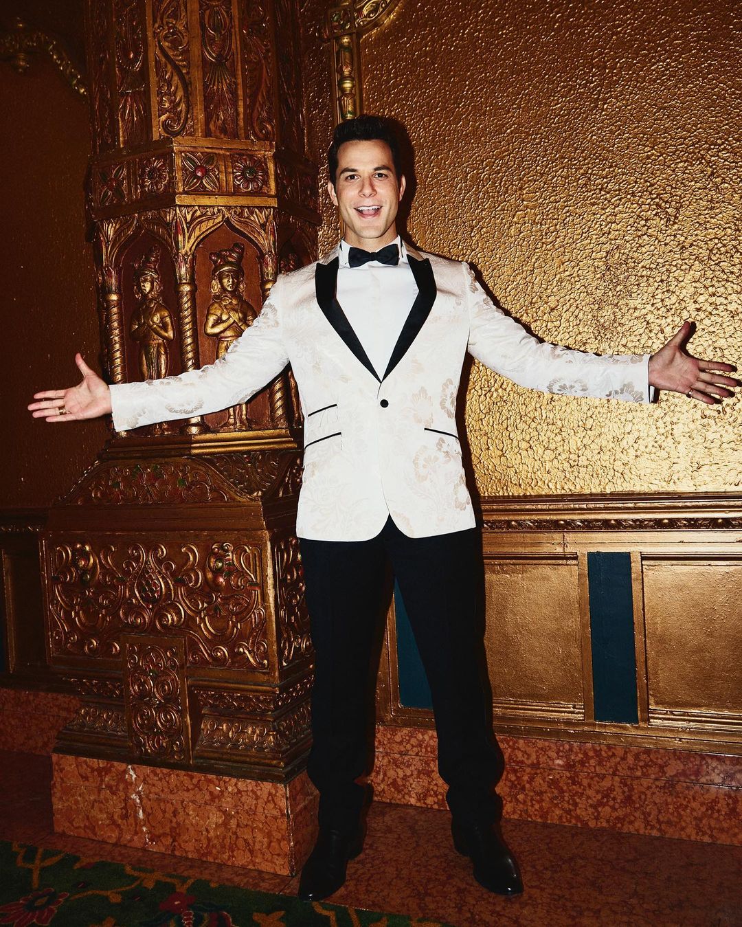 Adult male in a cream colored tuxedo with black trim and a bow tie smiles with his arms out in a gold colored ornate interior.