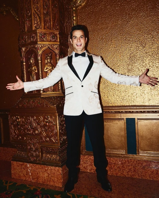 Adult male in a cream colored tuxedo with black trim and a bow tie smiles with his arms out in a gold colored ornate interior.