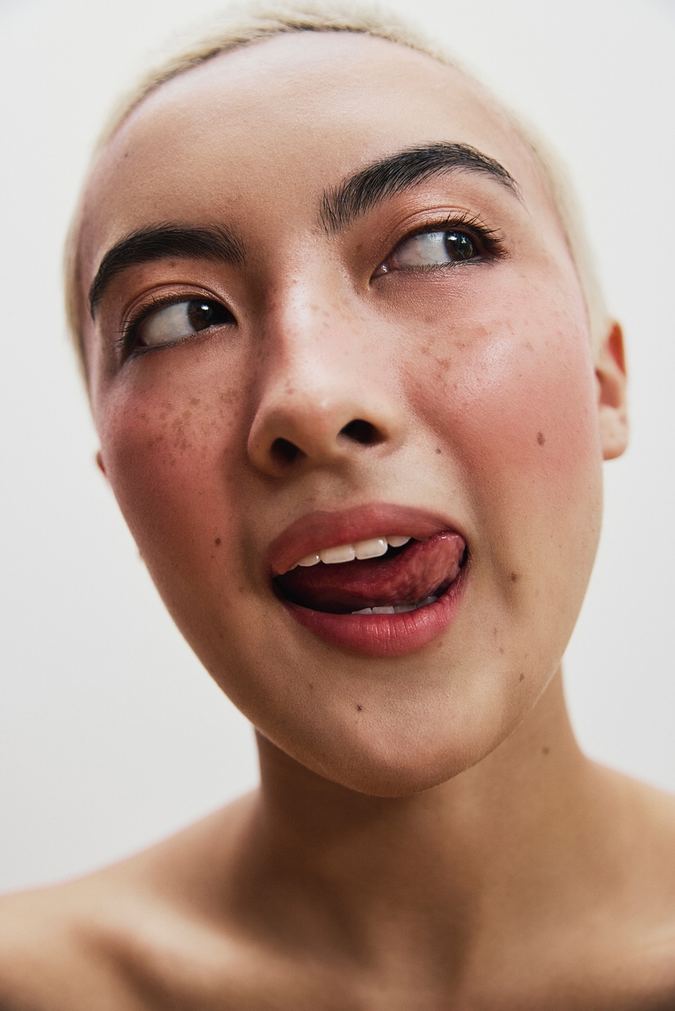 Close-up portrait of a young adult woman with freckles and short blonde hair, sticking her tongue out against a white background.