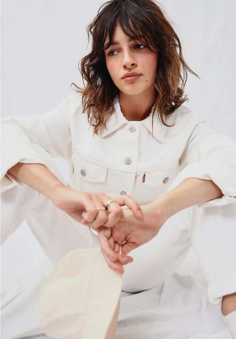 A young woman with brown hair poses indoors, wearing a white denim jacket and jeans, holding a cream-colored hat in a bright photo studio.