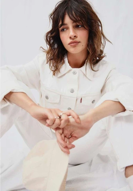 A young woman with brown hair poses indoors, wearing a white denim jacket and jeans, holding a cream-colored hat in a bright photo studio.