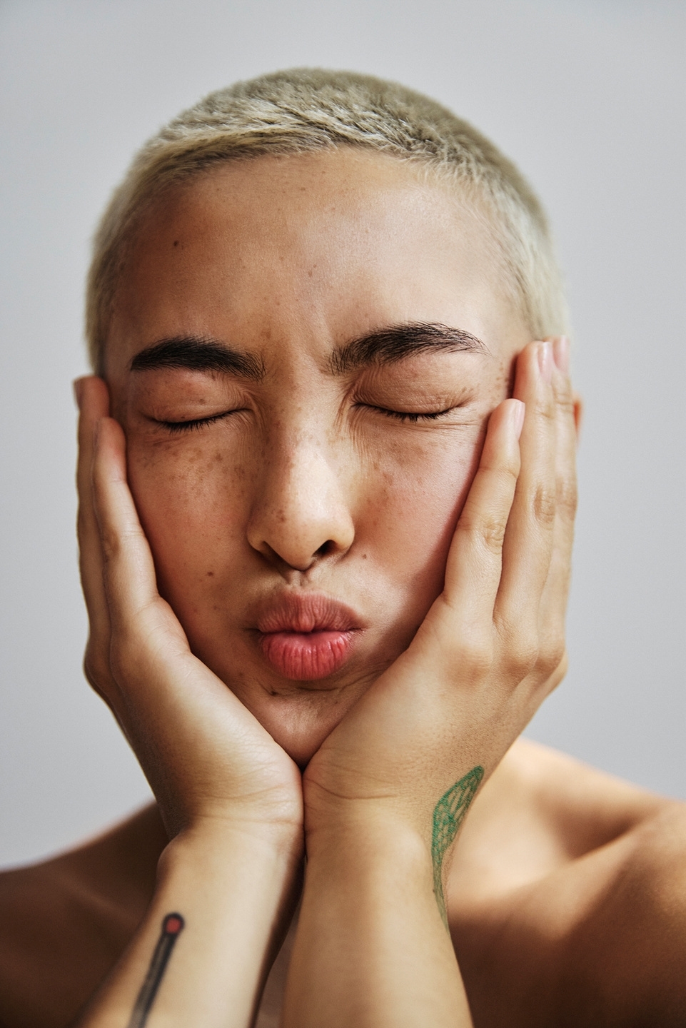 Portrait of a young adult woman with a buzzcut and light skin tone, her eyes closed, holding her face in her hands, against a white backdrop.