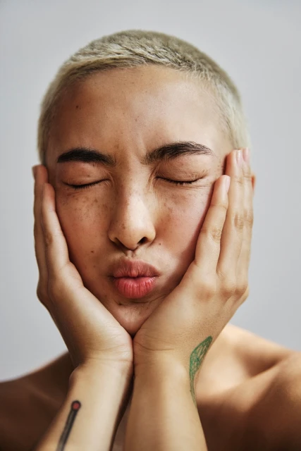 Portrait of a young adult woman with a buzzcut and light skin tone, her eyes closed, holding her face in her hands, against a white backdrop.