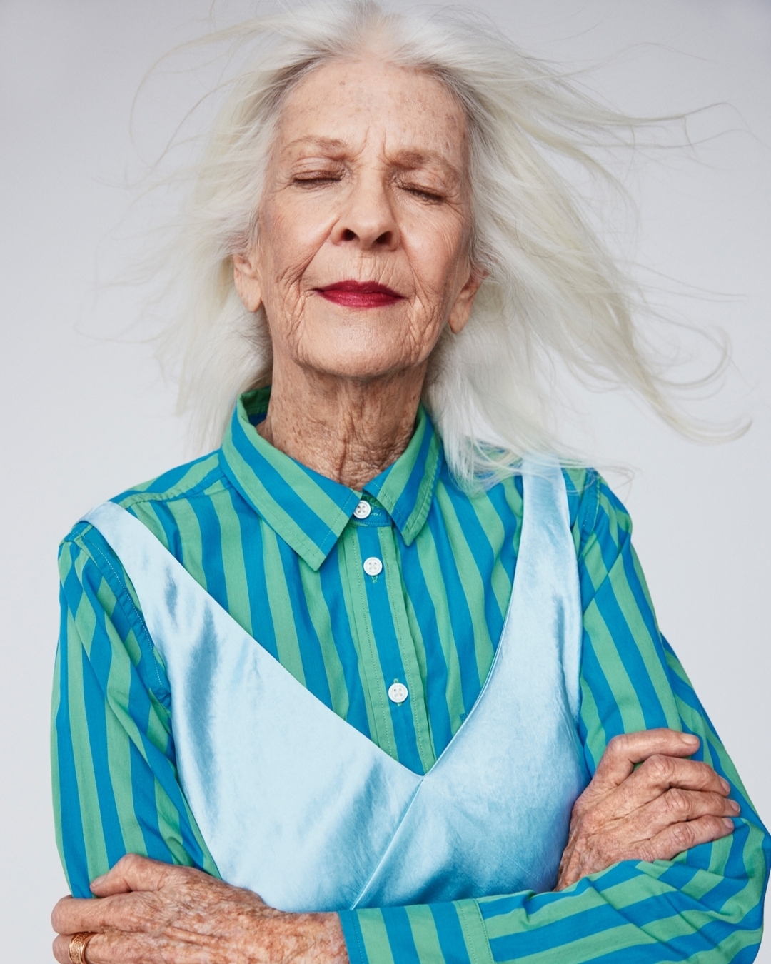 Portrait of a senior woman with gray hair, eyes closed, wearing a striped shirt and light blue dress, captured in a studio setting.