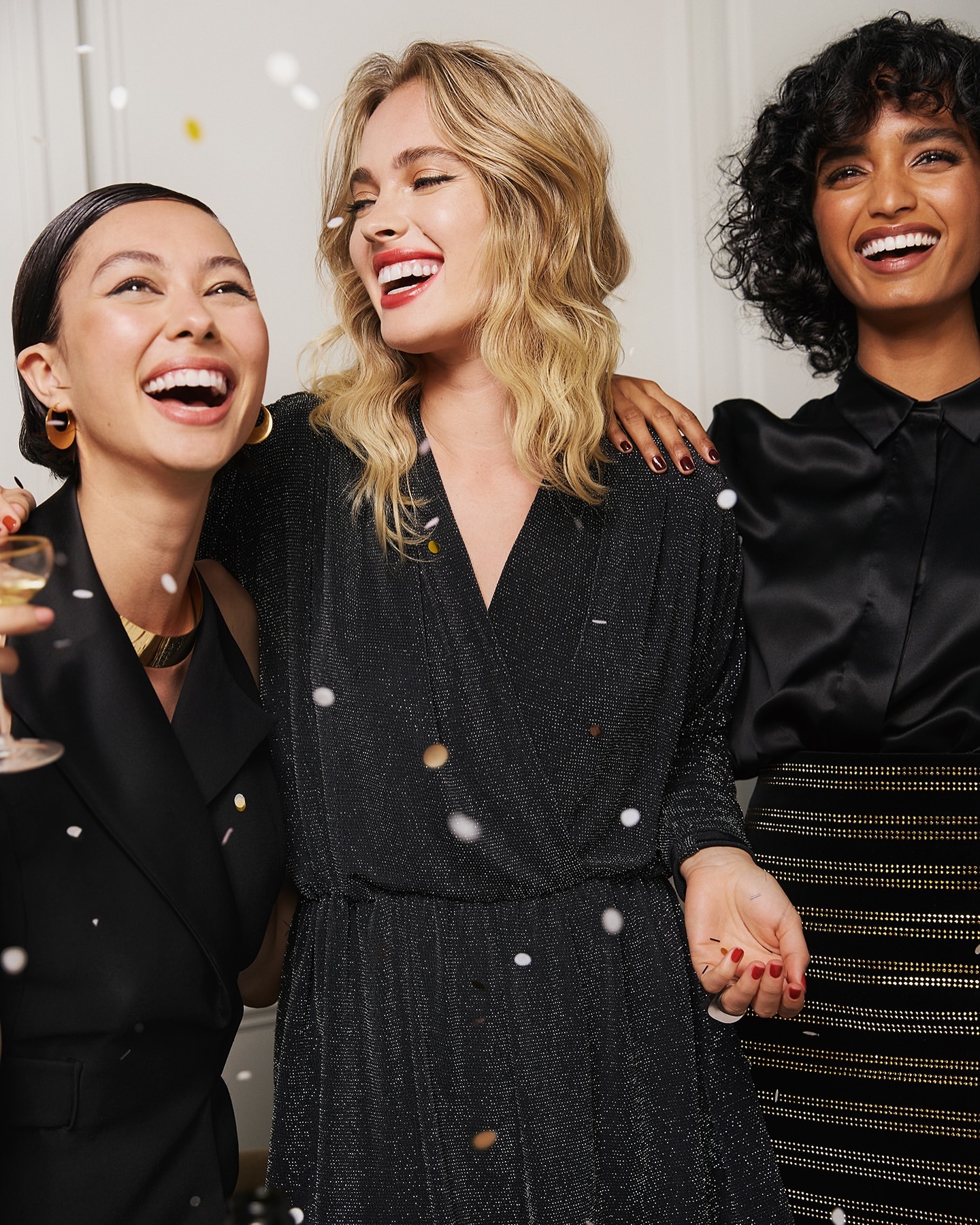 Three smiling multi-ethnic women in festive dresses celebrate with confetti. Indoor party or fashion editorial concept.