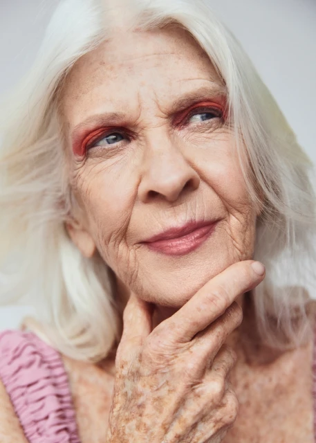 Close-up portrait of a smiling mature woman with white hair and red eyeshadow, resting her chin on her hand in a studio setting.