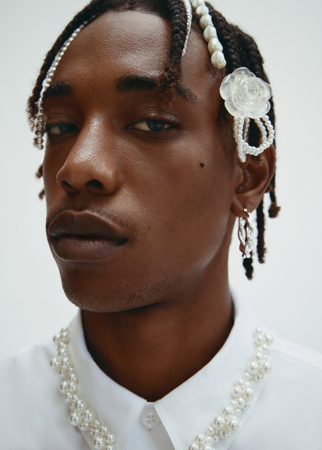 Close-up of a stylish Black man with beaded braids, wearing a white collared shirt embellished with pearl beads, against a white backdrop.