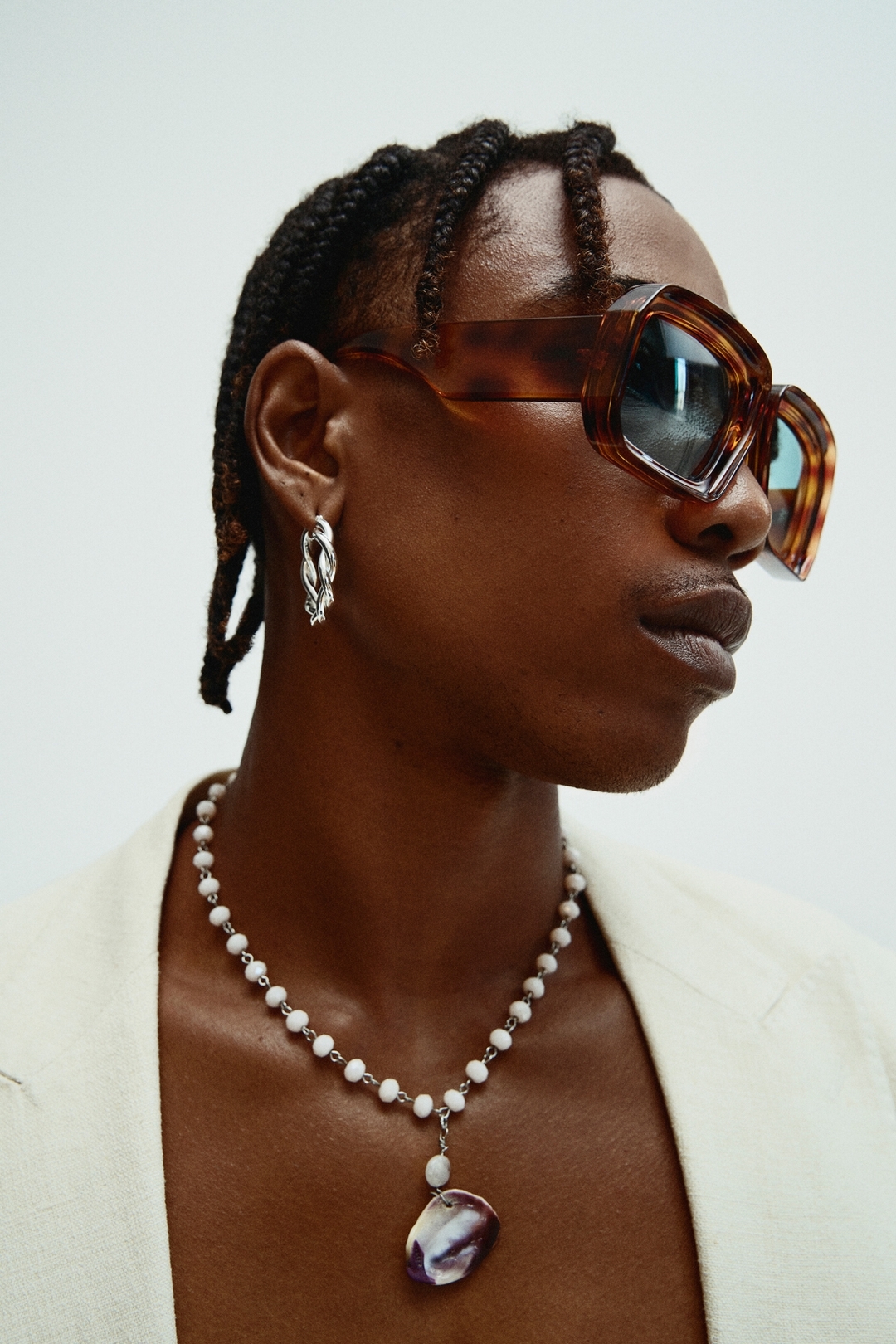 Portrait of a Black man wearing oversized tortoiseshell sunglasses, silver hoop earrings, and a white beaded necklace by Heather Moore.