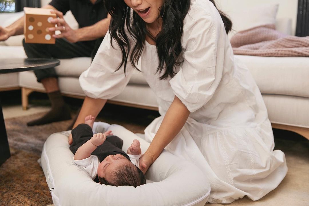 Mother looks on at her infant in a baby lounger in a warm interior, while an adult male holds a polka dot patterned cube in the background.