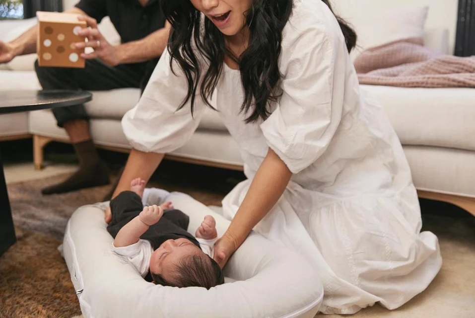 Mother looks on at her infant in a baby lounger in a warm interior, while an adult male holds a polka dot patterned cube in the background.