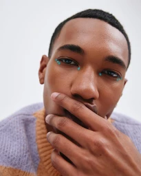 Close-up portrait of a Black man with turquoise dots of makeup around his eyes, wearing a textured knit sweater, against a plain background.
