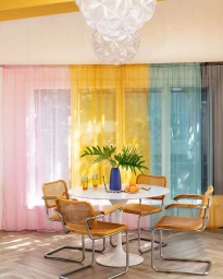 Round white dining table surrounded by four cane chairs in front of colorful sheer curtains in New York