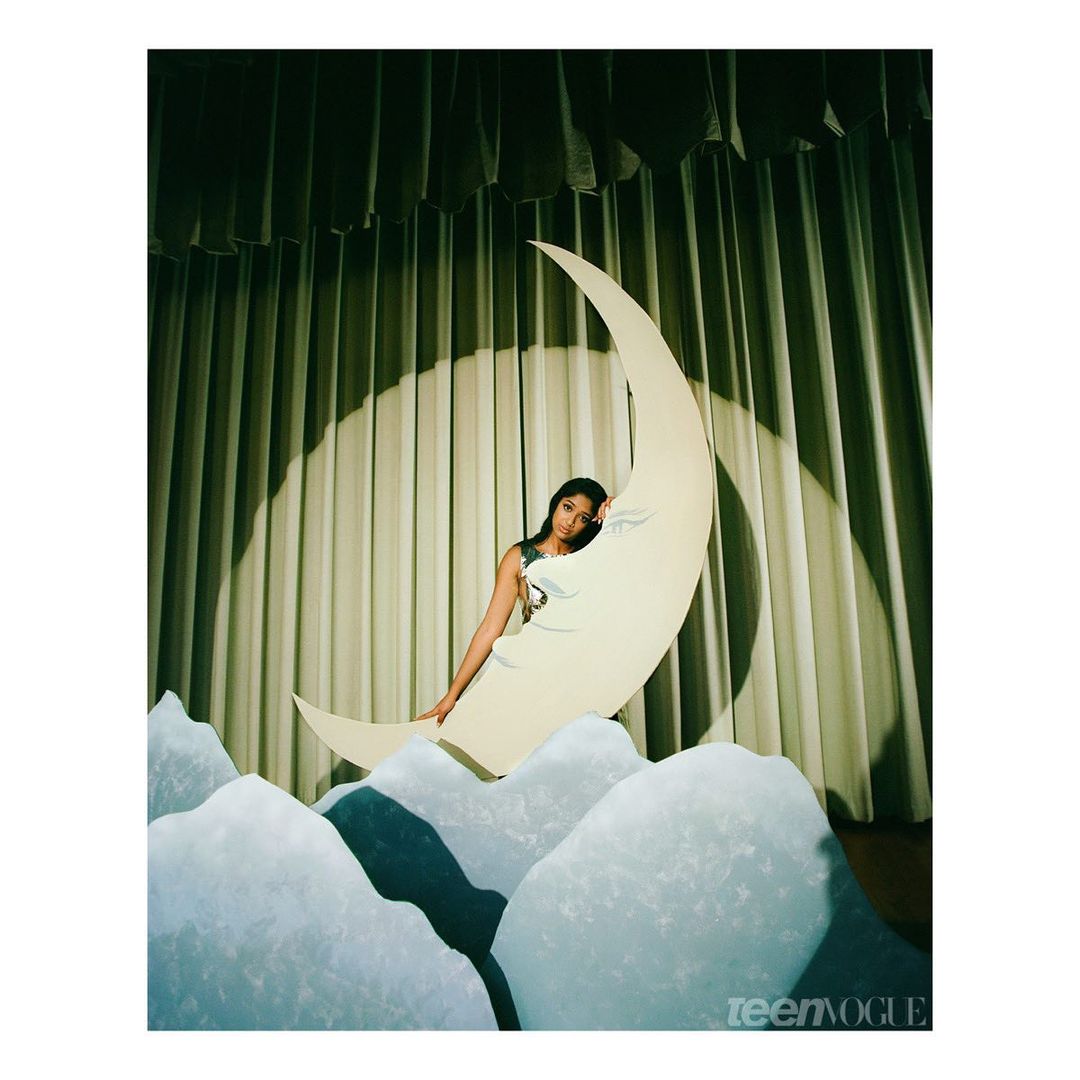 A young woman sits on a crescent moon prop in front of a stage curtain with cloud-like props in a colorful studio setting.