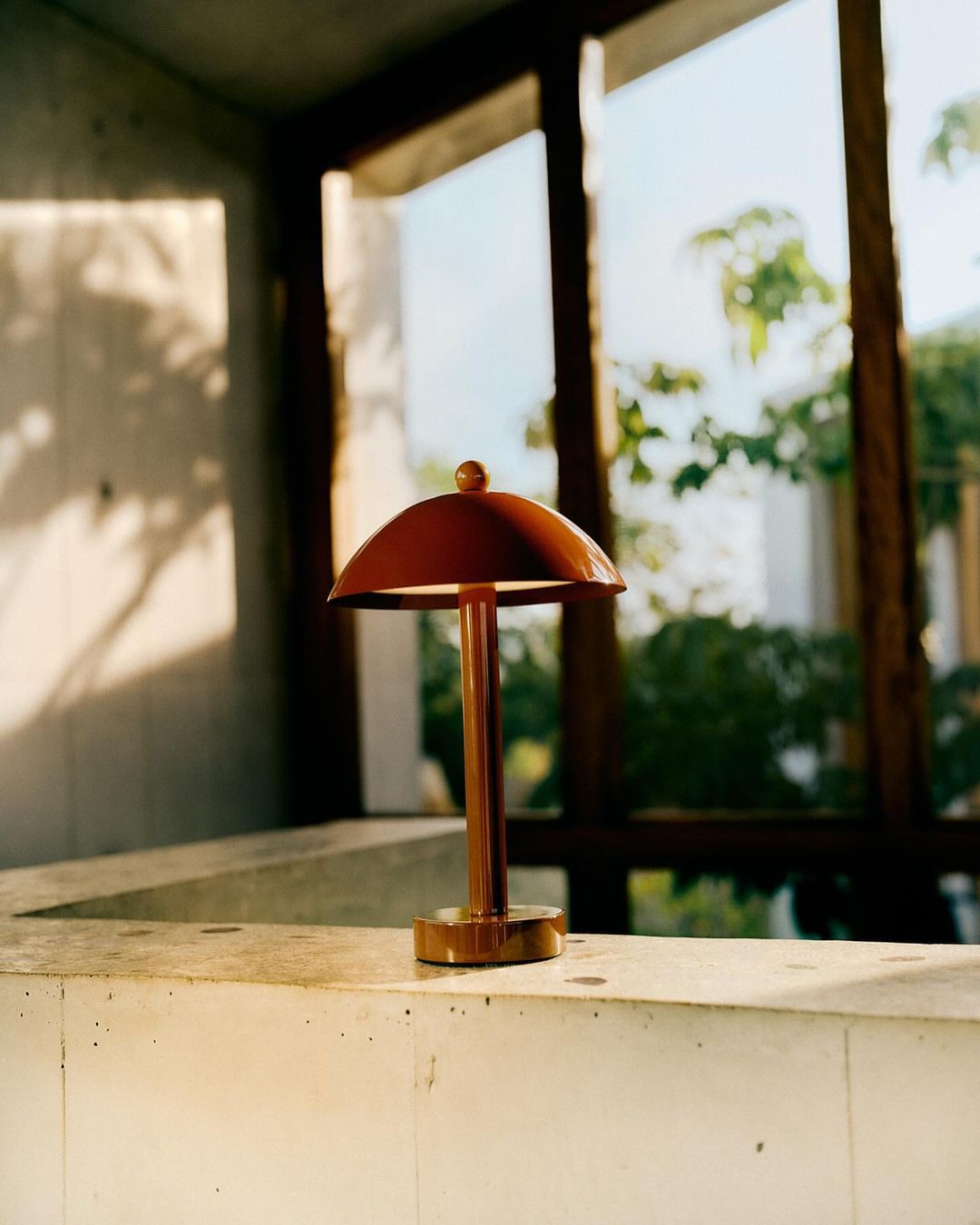 A modern copper-colored table lamp with a domed shade sits on a ledge in front of a window, casting soft light in an interior setting.