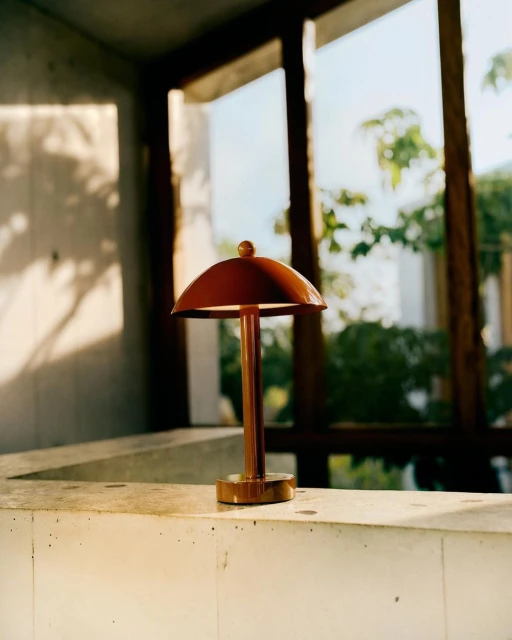 A modern copper-colored table lamp with a domed shade sits on a ledge in front of a window, casting soft light in an interior setting.