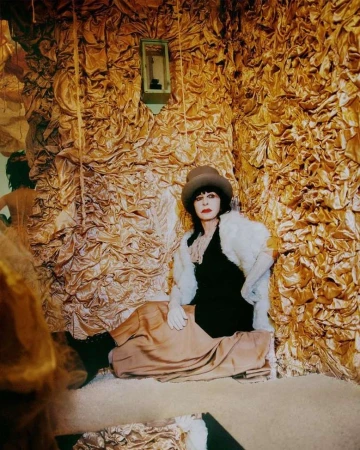 Person wearing a top hat and white fur stole seated against a textured gold fabric wall.