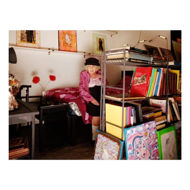 Elderly woman poses indoors near a bookshelf laden with books and paintings, wearing a pink jacket and a black hat.