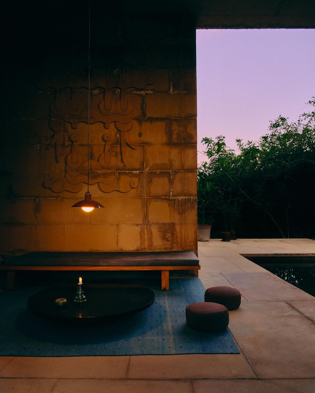 Outdoor patio at dusk features a stone bench, a round table with a candle, and garden views, illuminated by a hanging pendant light.
