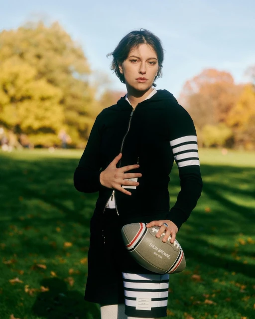 Adult female posing outdoors on grass, wearing black hooded jacket and skirt with white stripes, holding a football.