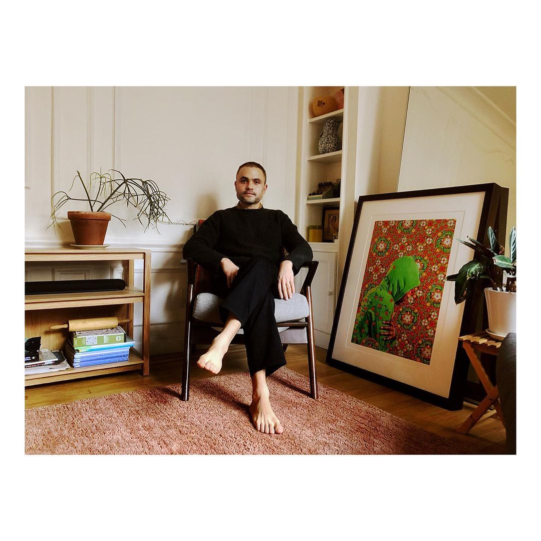 Indoor portrait of a barefoot man sitting in mid-century chair, next to framed art, plant, and cabinet in bright, natural light.