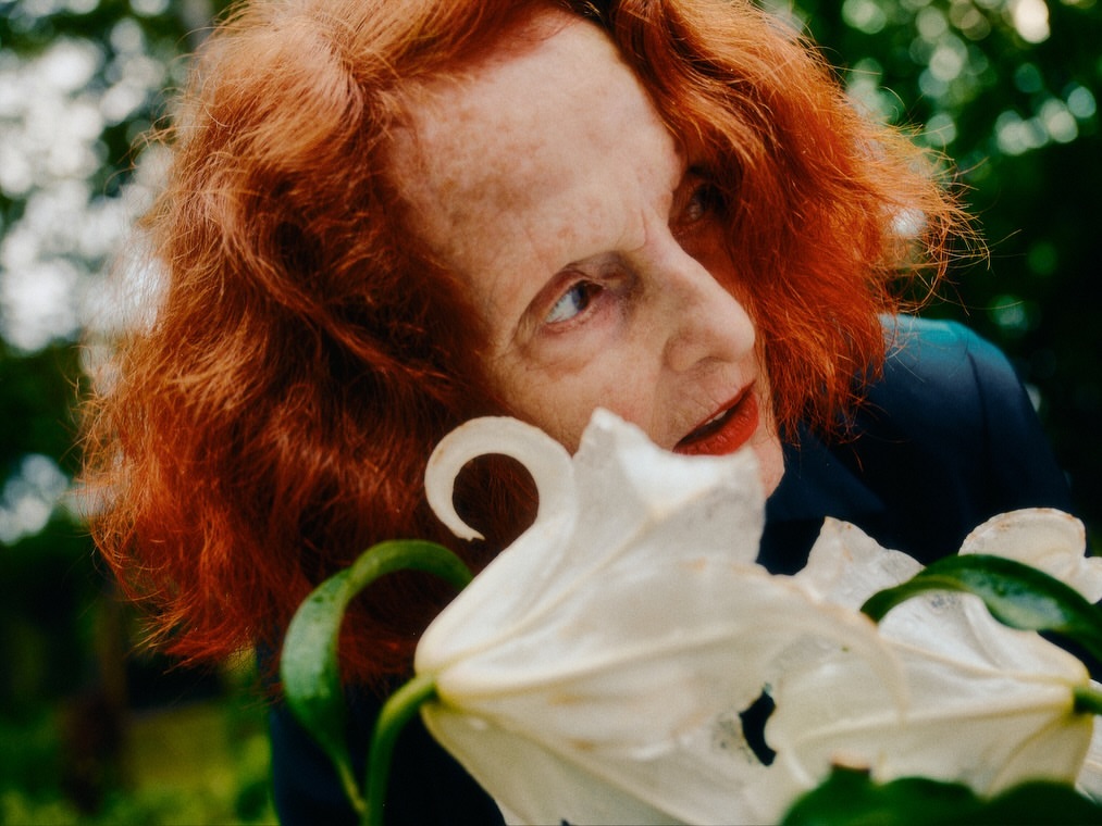 Close-up portrait of a woman with red hair and red lipstick standing outdoors next to a white lily in a garden setting.